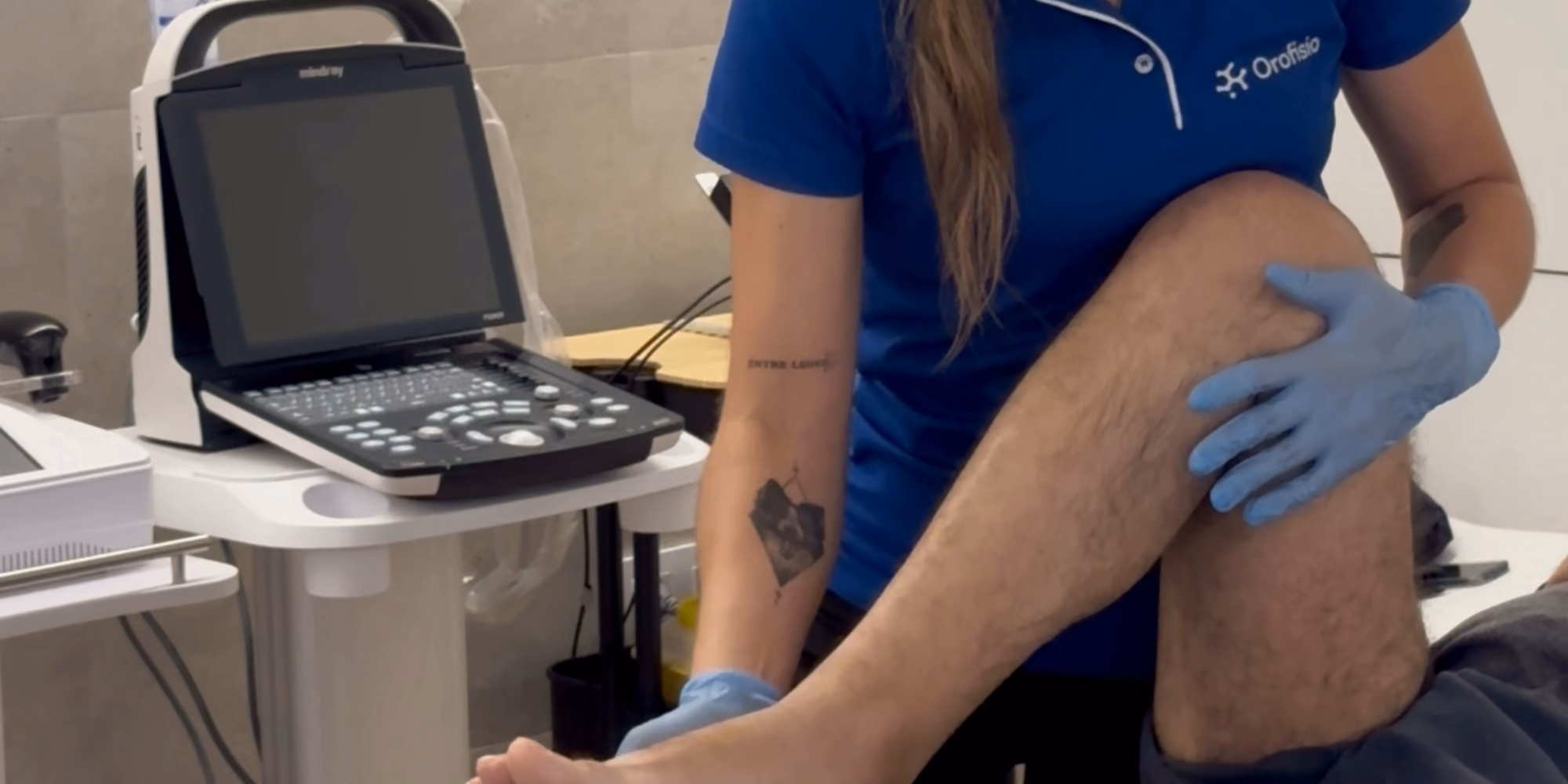 Una fisioterapeuta con el polo de Orofisio explora la rodilla de un paciente con la sonda del ecógrafo, con la pantalla del equipo encendida a su lado